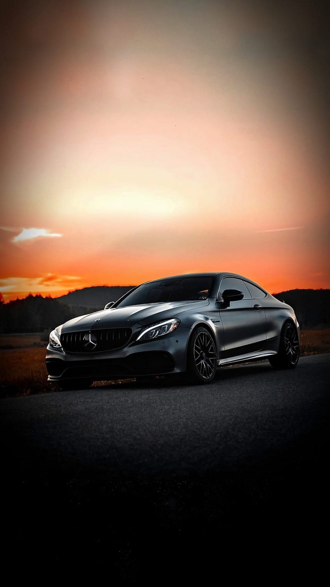 Dark Mercedes-Benz coupe parked on asphalt under an orange sunset sky with silhouetted trees in the distance.