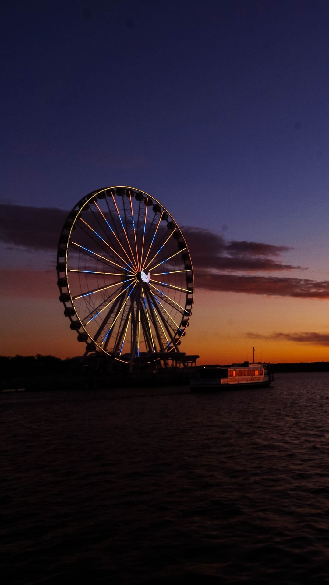 Ferris wheel with yellow and blue lights stands by a river under an orange and indigo sunset sky with a boat.