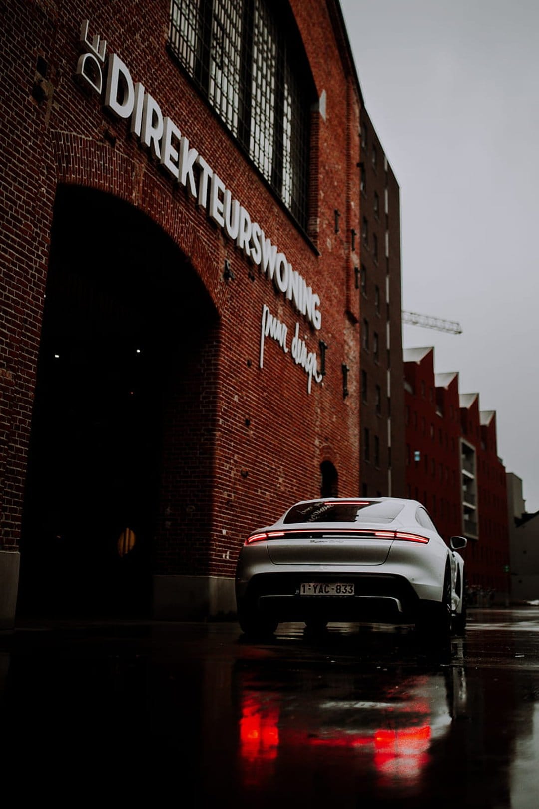 Silver Porsche Taycan with red taillights parked on a wet street in front of a historic red brick building.