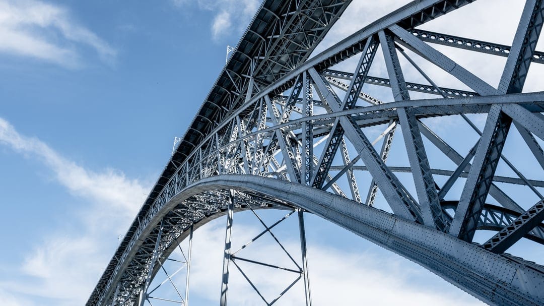 Steel arched bridge girders rise into a vibrant blue sky with wispy clouds from a dramatic low-angle perspective.