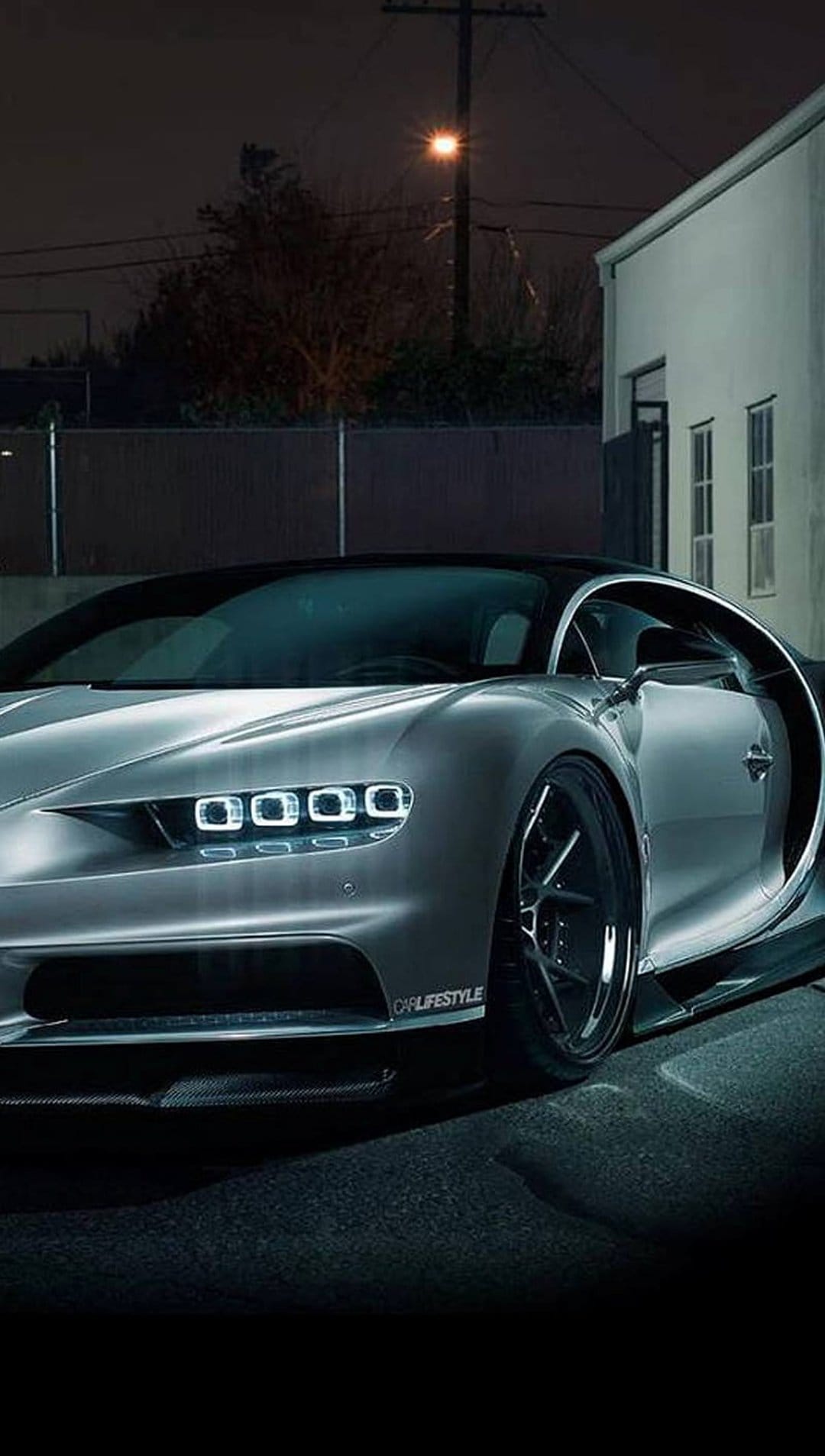 Silver Bugatti Chiron hypercar parked on asphalt at night with glowing quad LED headlights and a black roof.
