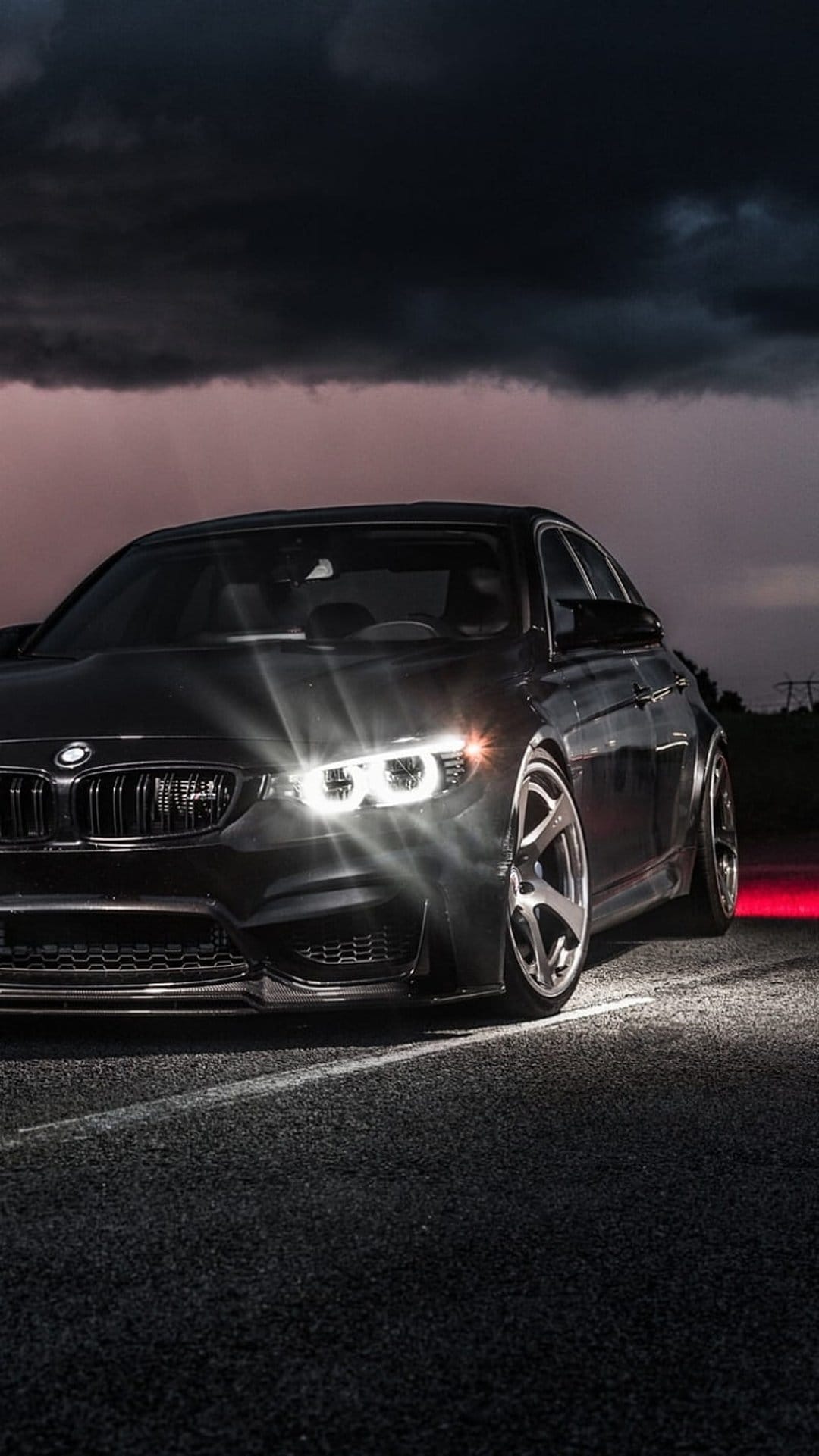 Black BMW M4 sports coupe with glowing LED headlights parked on a damp road under a stormy twilight sky.