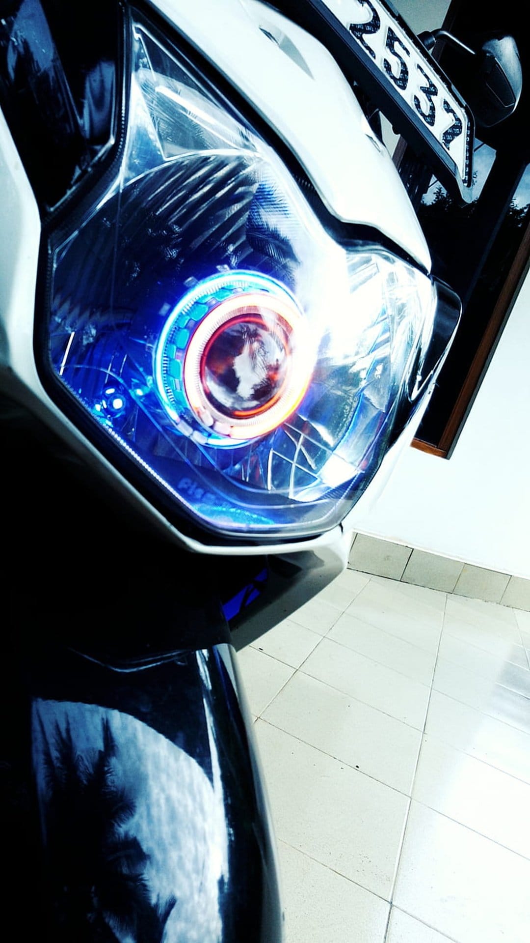 Motorcycle headlight with a glowing blue LED halo ring and chrome fairing reflecting tropical palm trees.