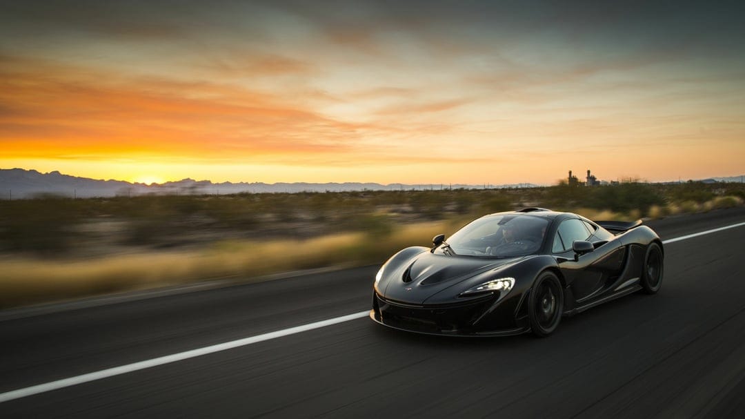 Black McLaren P1 hypercar driving fast on a desert highway during a vibrant orange and yellow golden hour sunset.