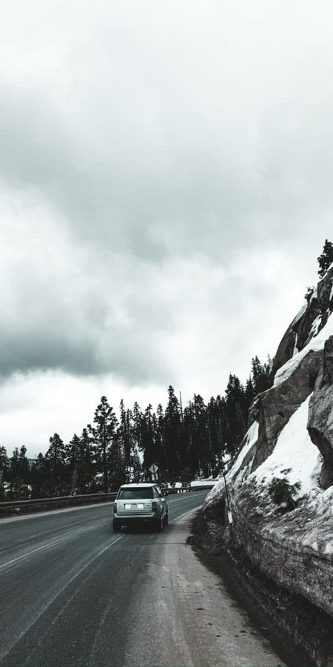 Silver SUV driving on a winding wet road between snowy rock walls and dense pine trees under a cloudy sky.