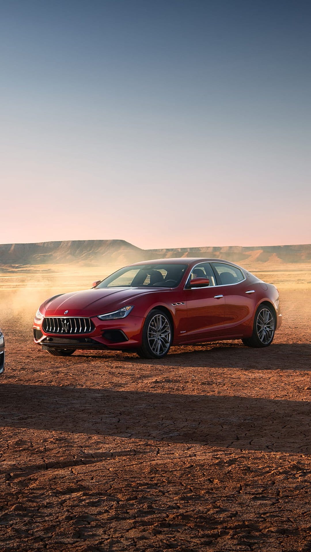 Red Maserati Ghibli sedan driving fast through a sandy desert with dust clouds swirling around the wheels.