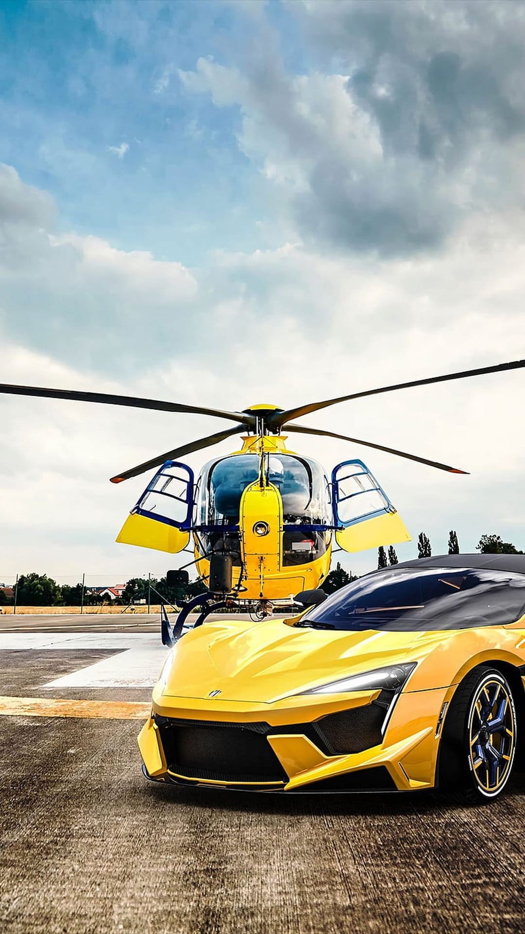 Yellow supercar and matching helicopter parked on an asphalt runway under a cloudy sky during the daytime.