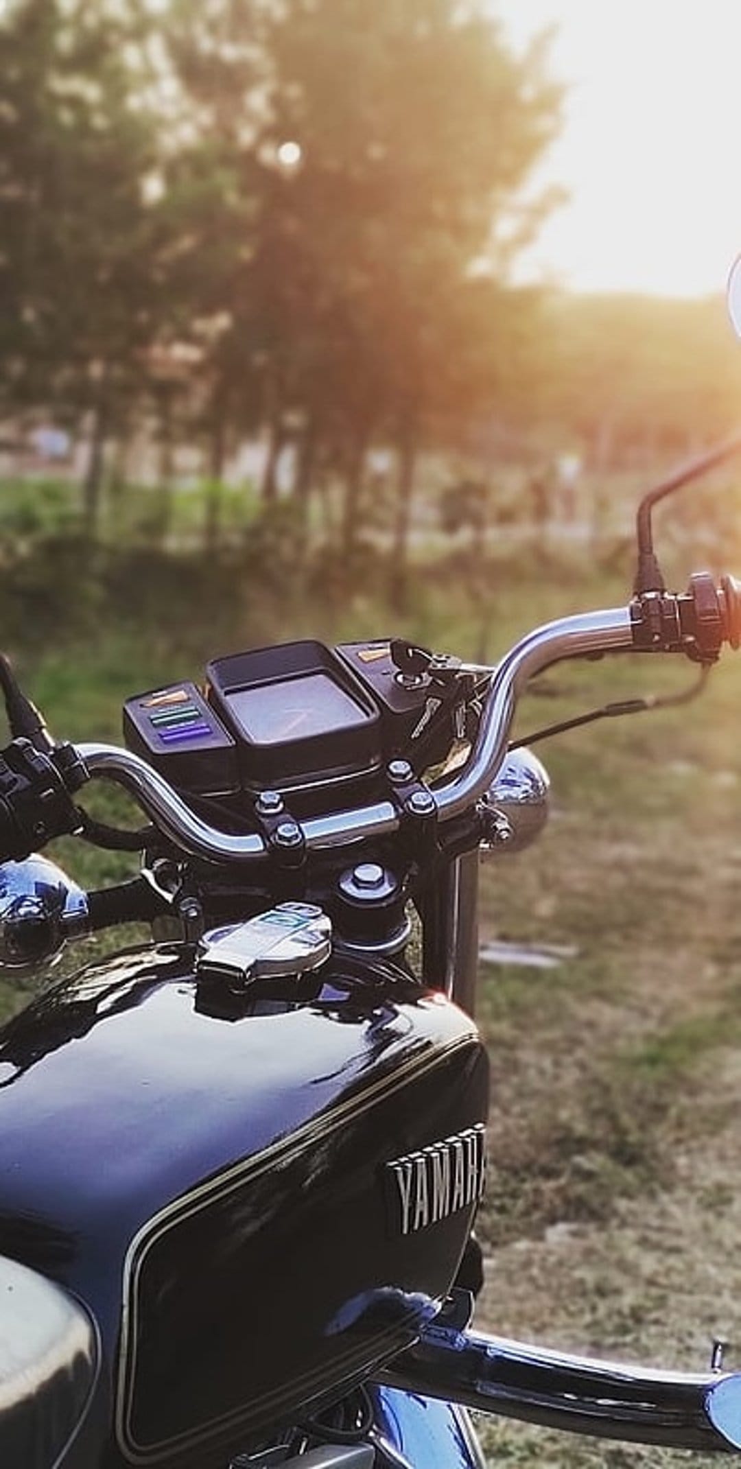 Yamaha motorcycle handlebars and fuel tank reflecting sunset light against a blurred green meadow backdrop.