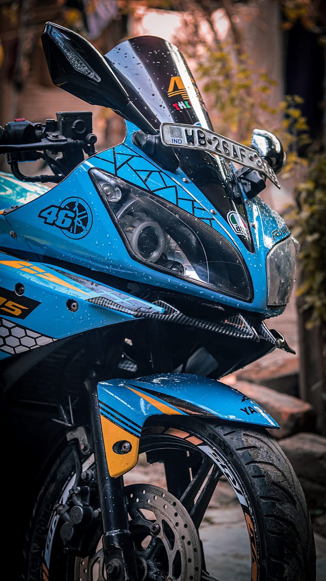 Blue Yamaha R15 motorcycle with rain droplets, carbon fiber accents, and 46 decals on a blurred background.