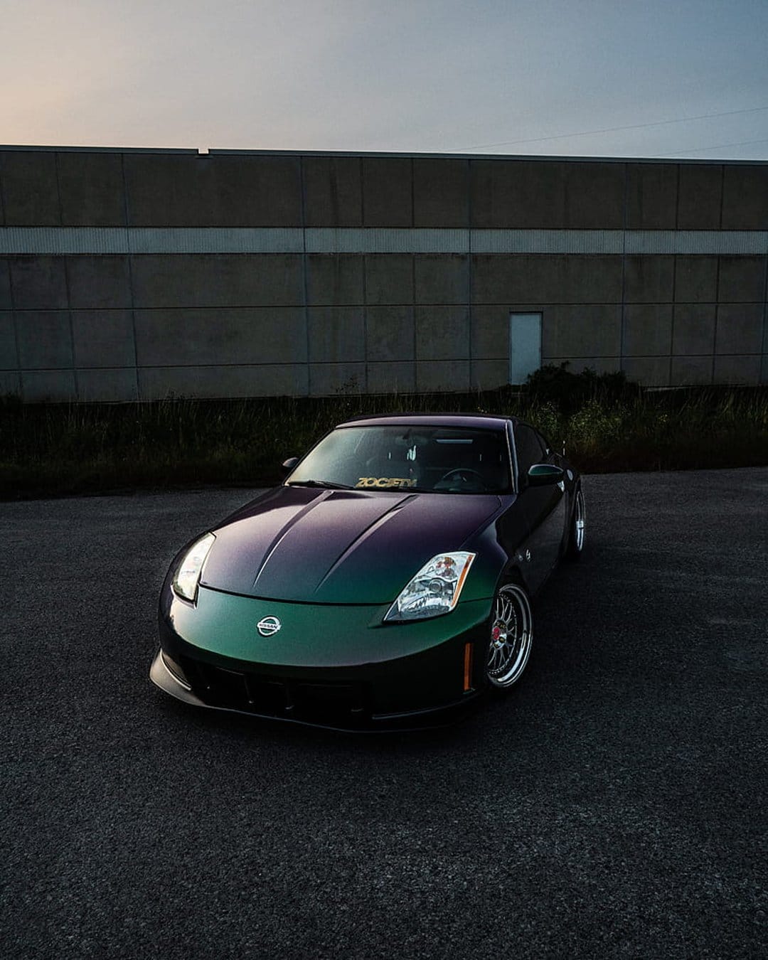 Nissan 350Z with green and purple iridescent paint parked on asphalt at dusk near an industrial backdrop.
