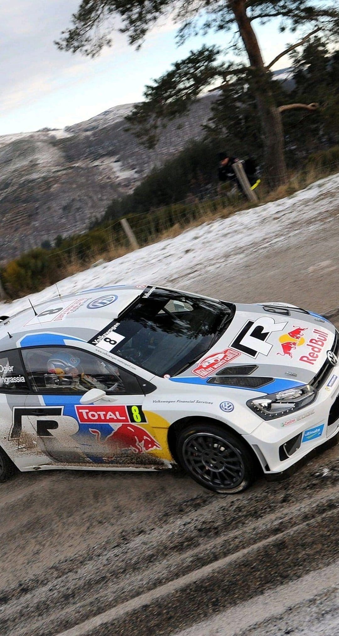 White Volkswagen rally car number 8 drifting on a snowy mountain road with evergreen trees and a clear blue sky