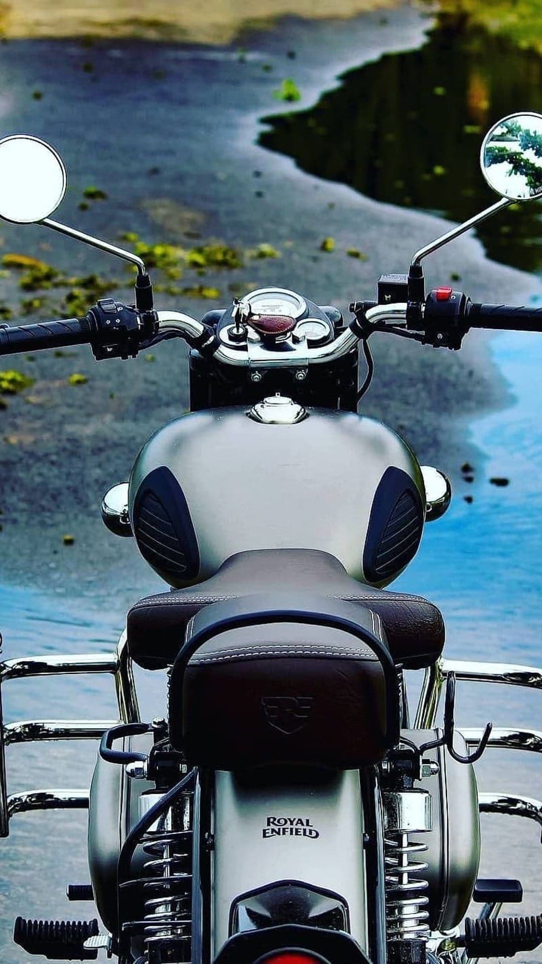 Royal Enfield motorcycle seat and handlebars featuring dark brown leather and chrome accents near reflective water.