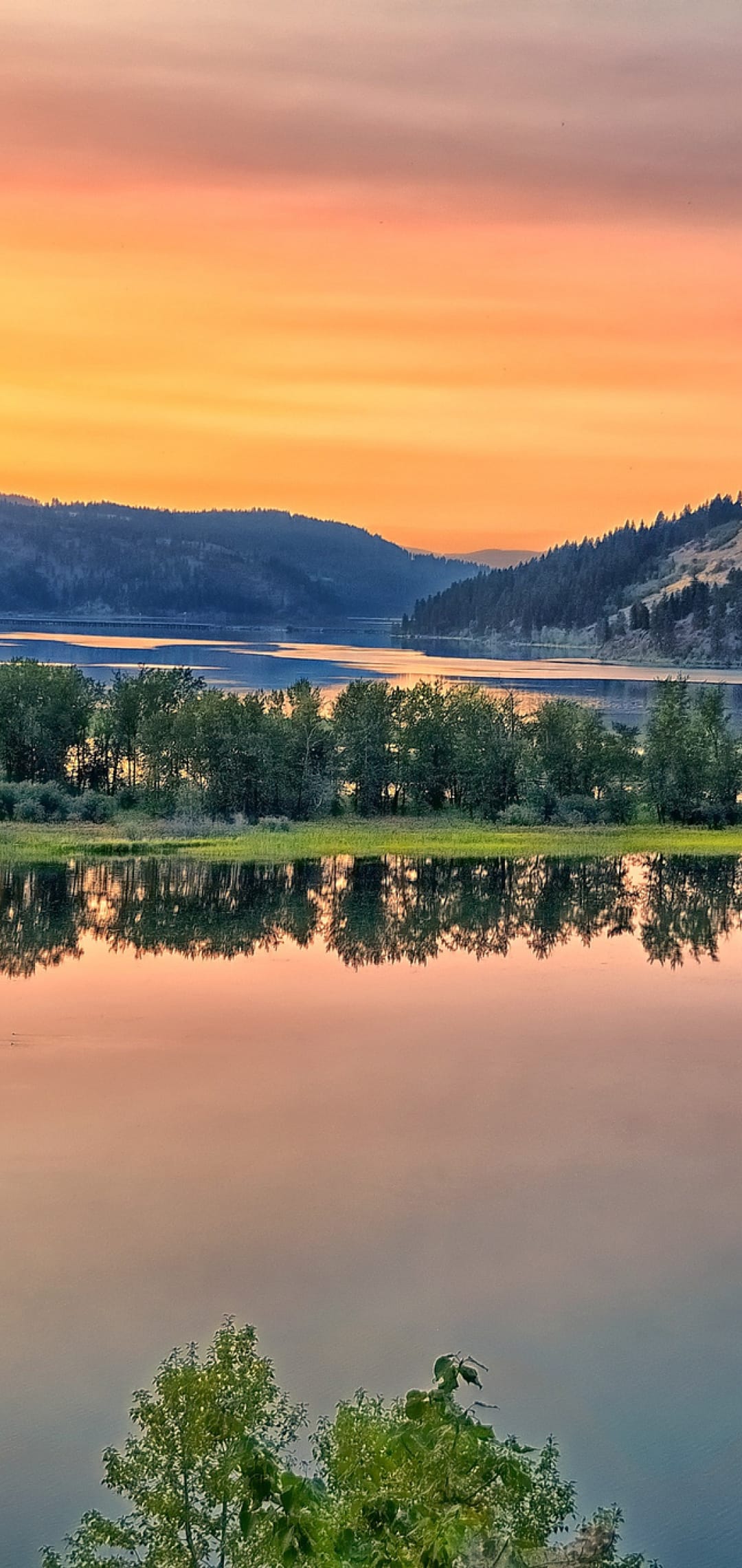 Calm river reflects a vibrant orange and blue sunset sky between silhouettes of trees and distant mountain peaks.