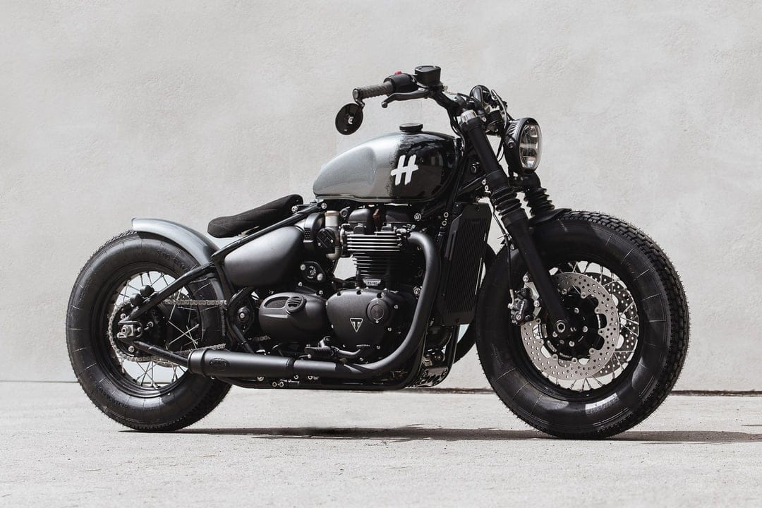 Custom black Triumph bobber motorcycle with grey tank parked against a minimalist white wall in bright light.