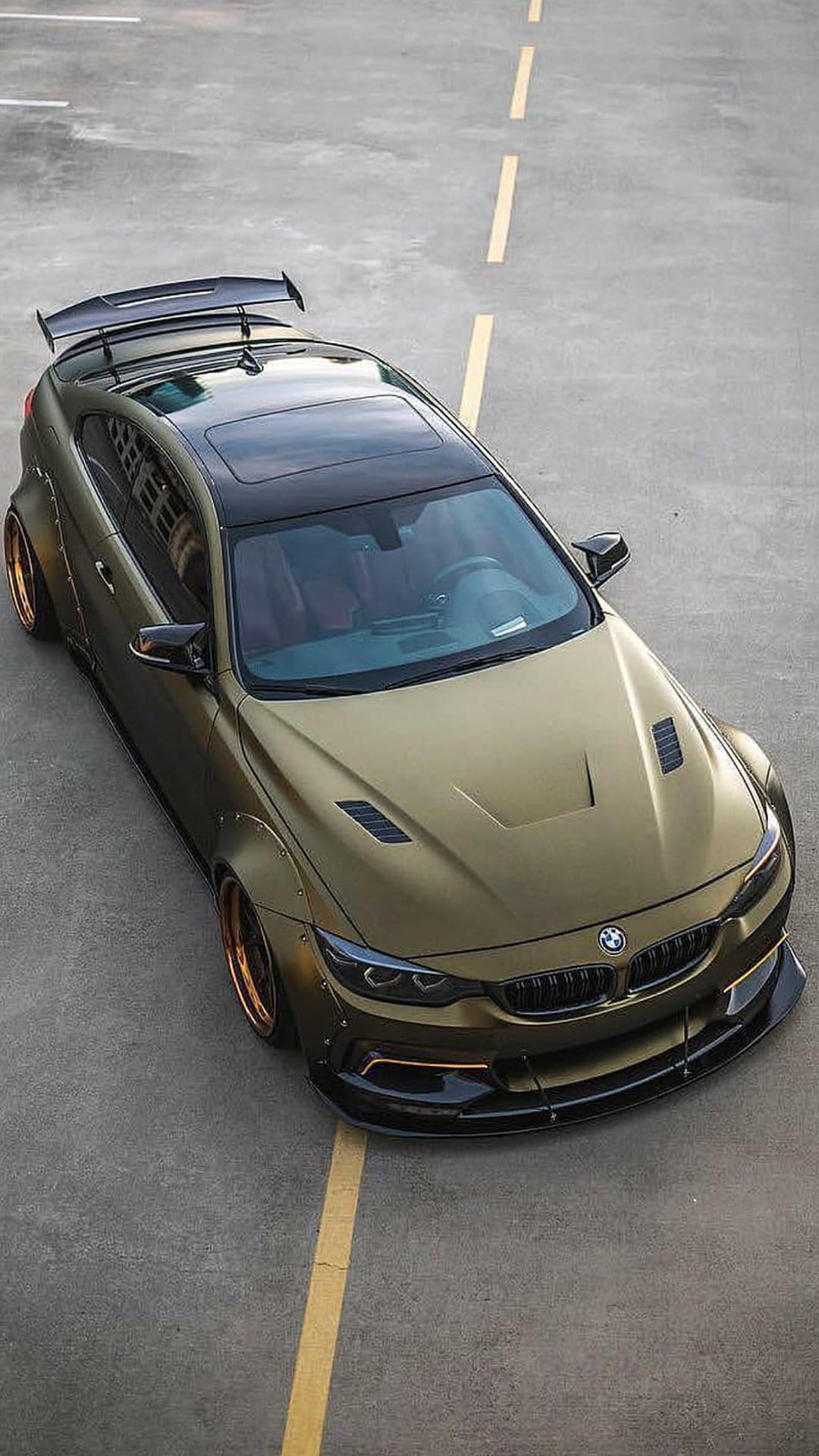 Gold BMW M4 coupe with widebody fenders, rear wing, and bronze wheels from an aerial drone perspective view.