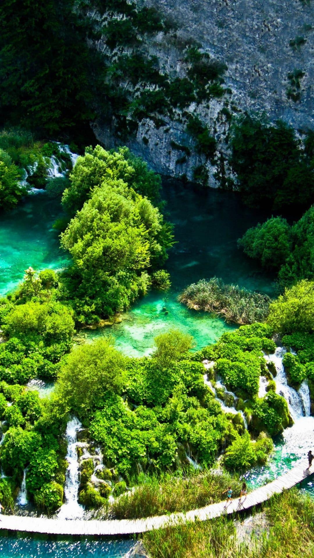 Turquoise Plitvice Lake waterfalls and a wooden boardwalk are viewed from above amidst lush green forest islands.
