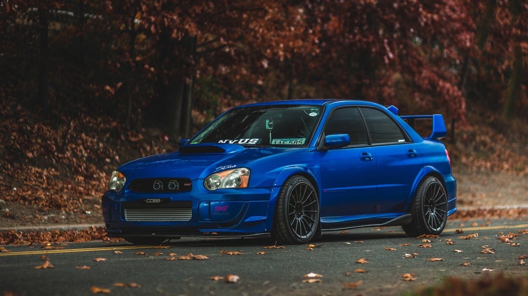 Blue modified Subaru WRX STI parked on an autumn road covered in fallen leaves with blurred trees.