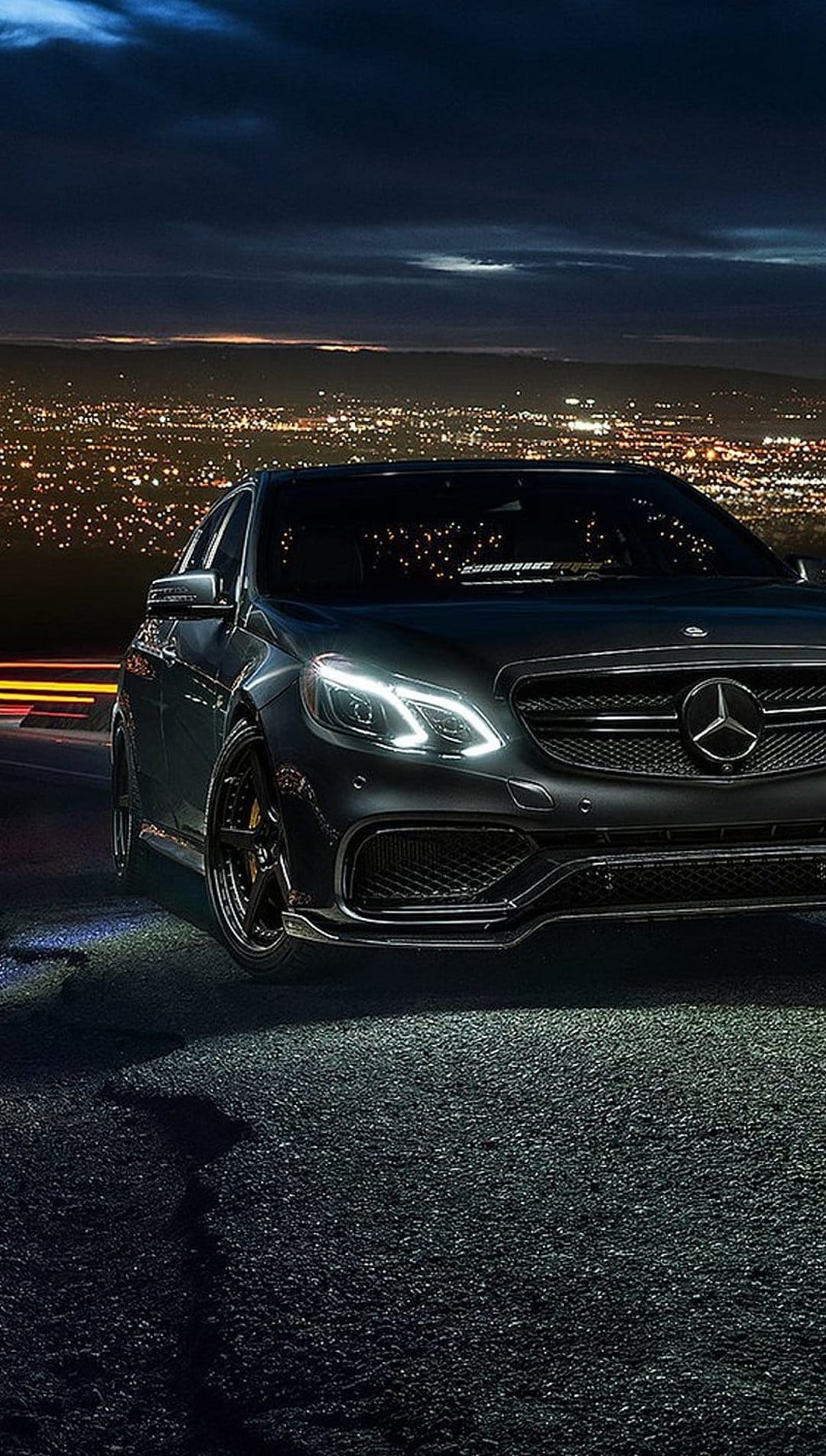 Black Mercedes E-Class sedan parked on a textured road with glowing headlights against a nighttime city skyline.