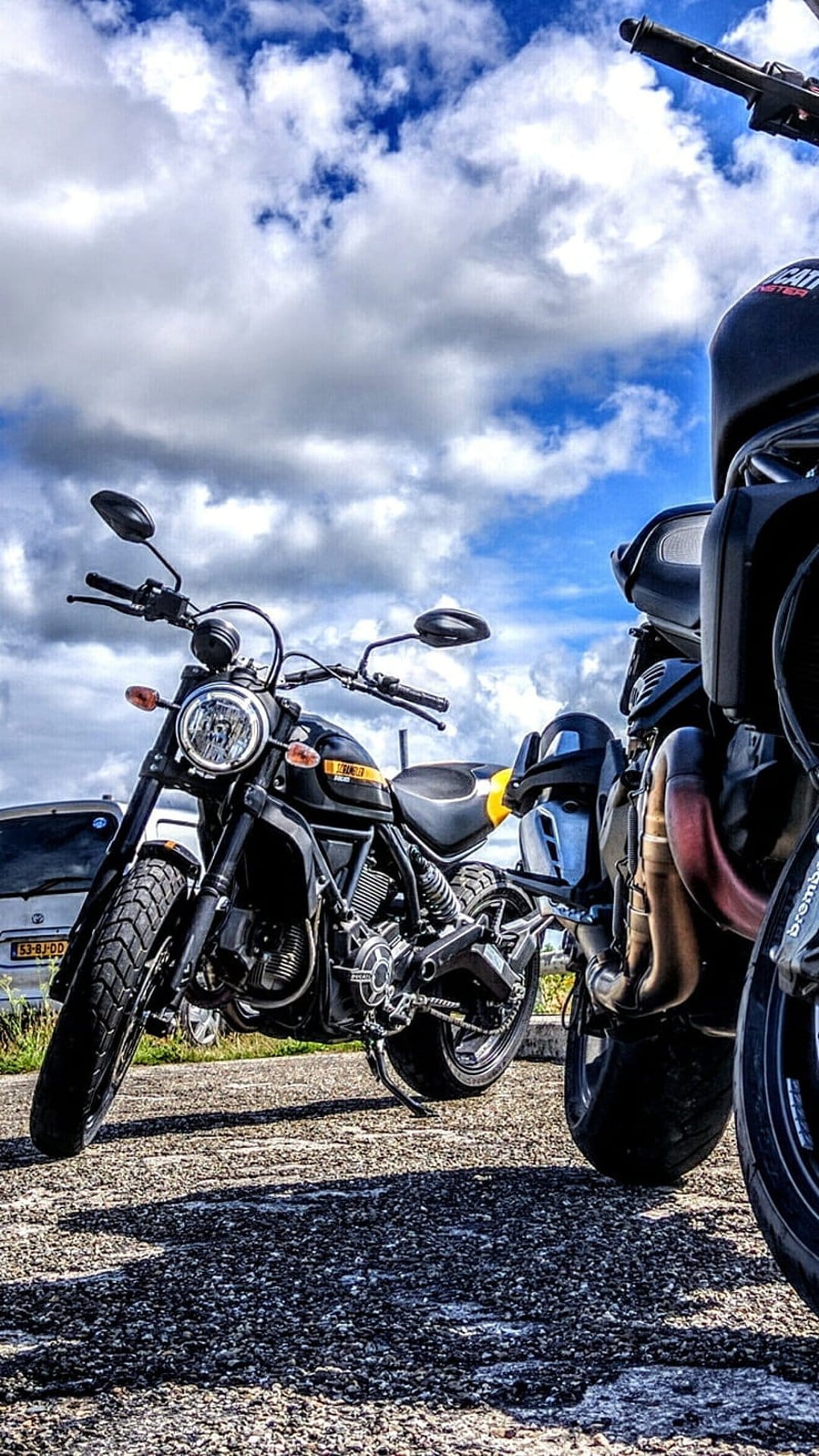 Two black motorcycles parked on a grey gravel surface against a backdrop of dark blue clouds in the daytime.