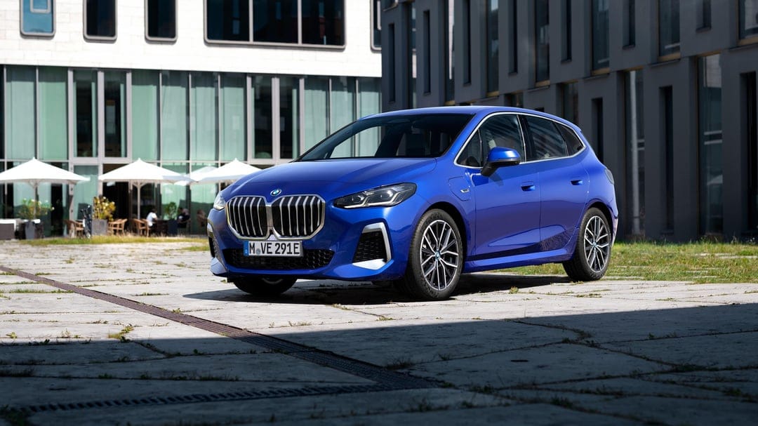 Blue BMW 2 Series Active Tourer parked on a sunny concrete plaza with modern glass buildings in the background.