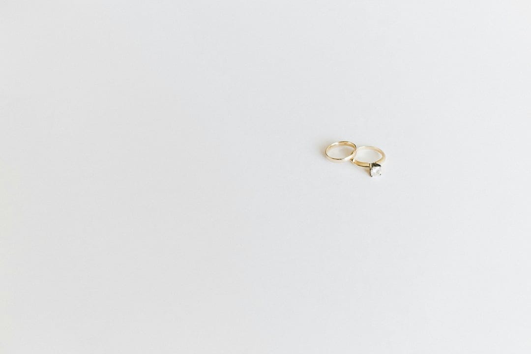 Two gold rings, including one with a small diamond, sit side-by-side on a seamless white studio background.