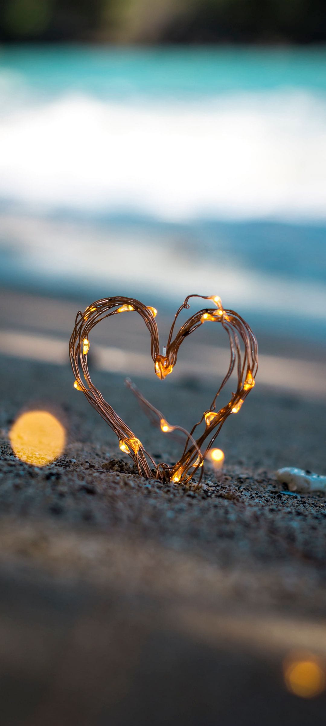 Heart shaped fairy lights glowing on wet sand at sunset with a blurred ocean background and golden horizon.