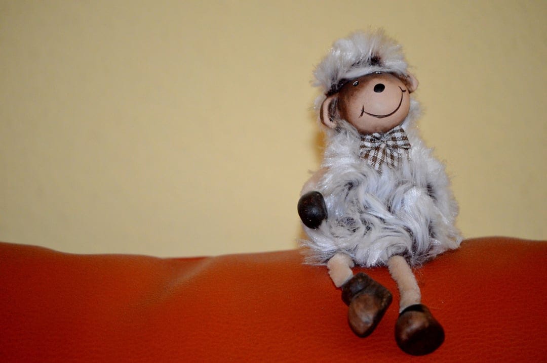 Fluffy grey sheep plush toy with a checkered bowtie sits on a vibrant orange sofa against a soft yellow wall.
