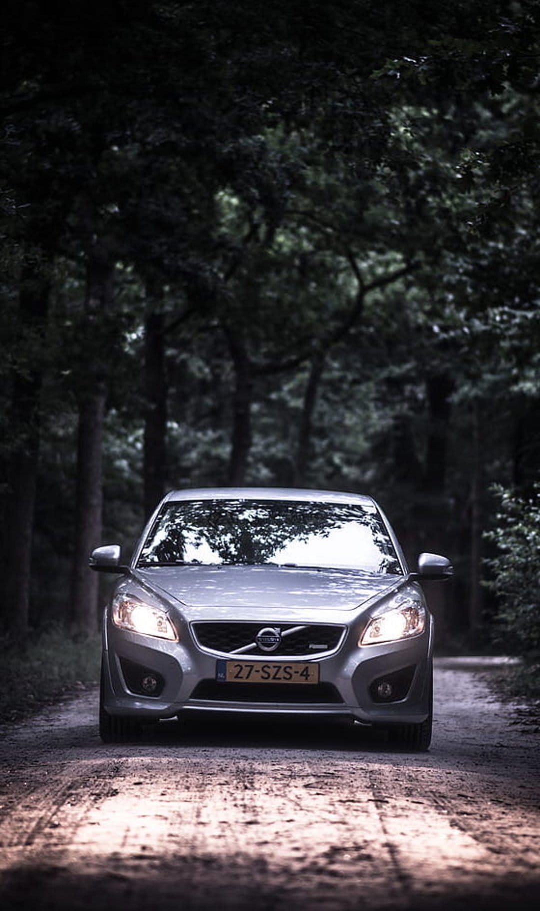 Silver Volvo C30 hatchback with lit headlights driving on a gravel road through a dense, dark pine forest.