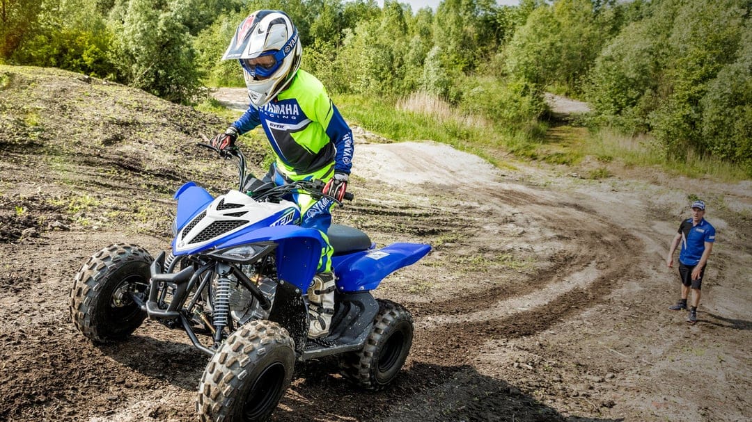 Child riding a blue Yamaha ATV on a dirt track through a forest setting with an instructor nearby.