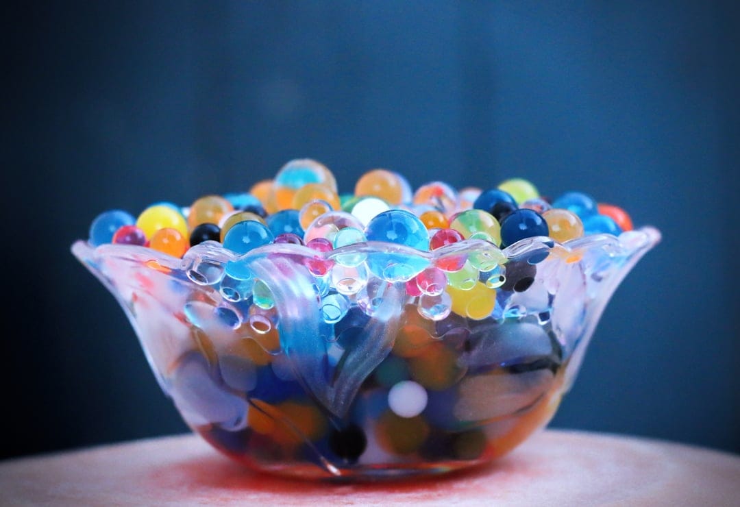 Multicolored translucent water beads inside a textured crystal bowl against a dark blue blurred background.
