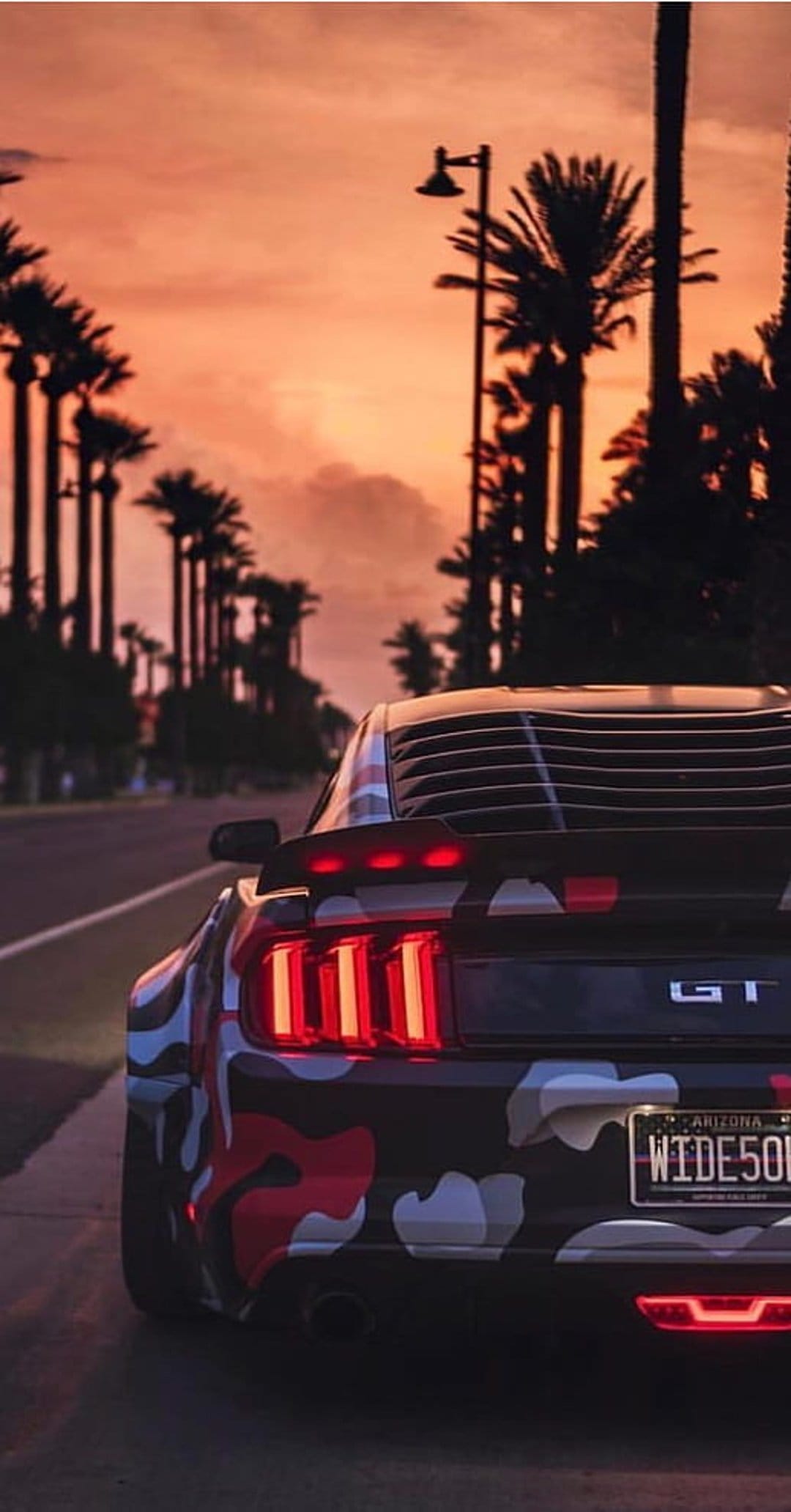 Ford Mustang GT with red camouflage wrap drives on a palm-lined road during a vibrant orange and pink sunset.