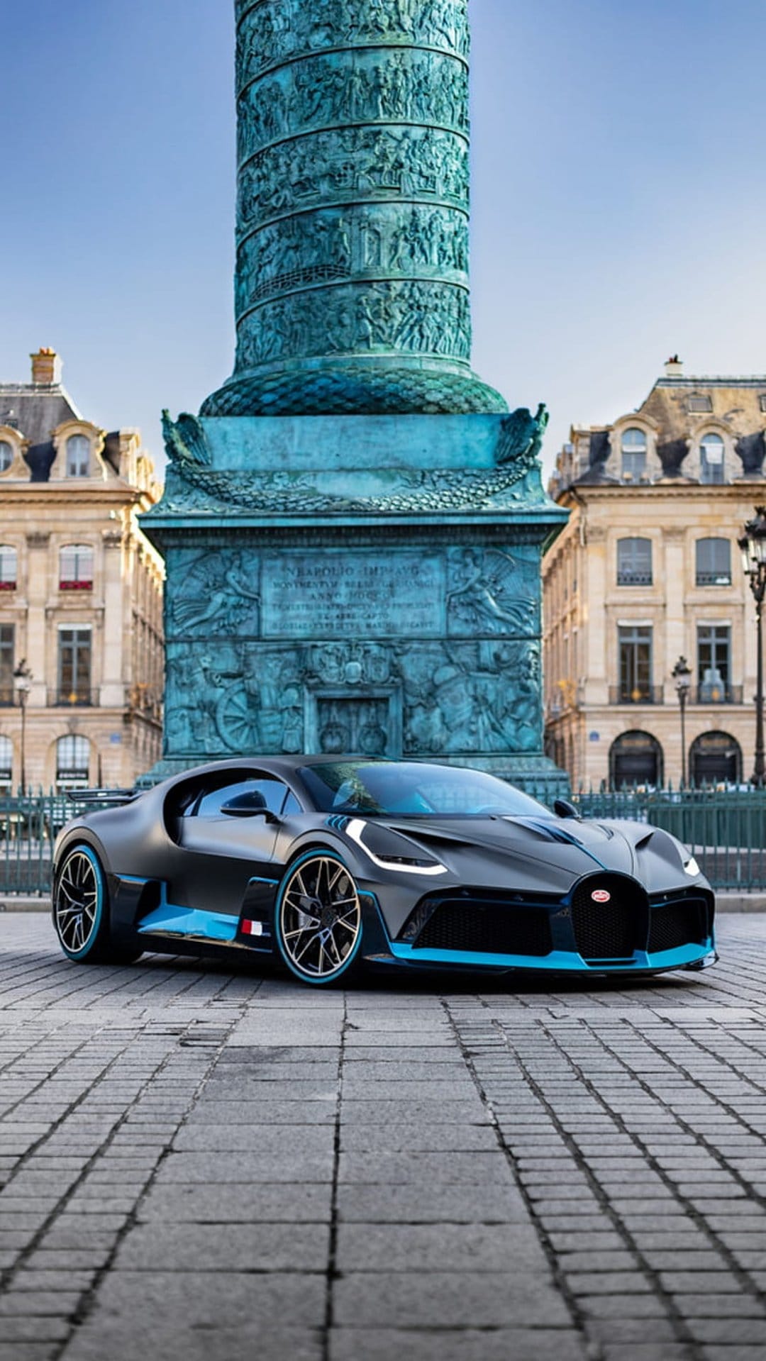 Black Bugatti Divo with blue trim parked on cobblestones before the Vendôme Column under a clear Parisian sky.