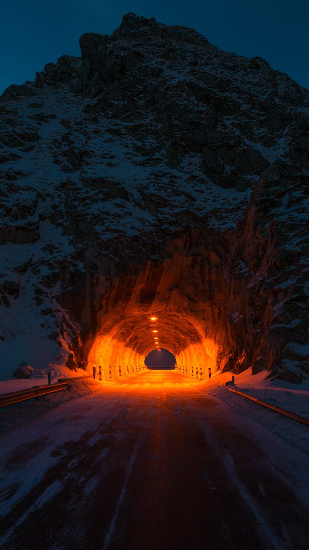 Orange-lit tunnel opening at the base of a jagged, snow-covered mountain peak under a dark blue twilight sky.