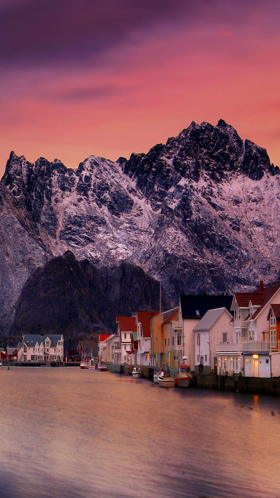 Snowy mountain peaks tower over a glowing Norwegian coastal village at dusk with reflections on calm water.