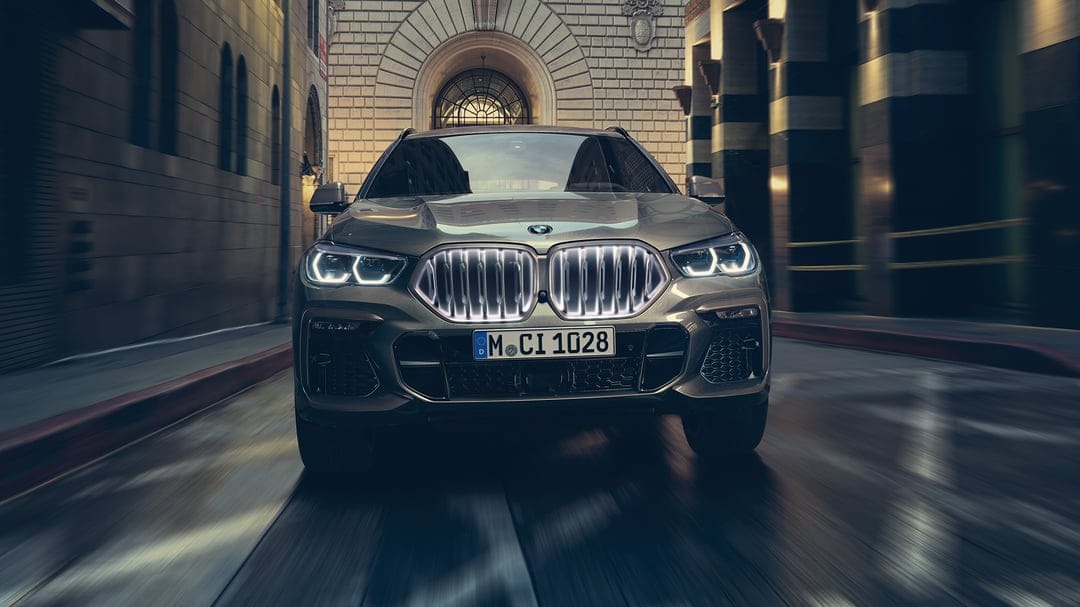 Front view of a grey BMW X6 with an illuminated grille driving on a city street with stone architecture.