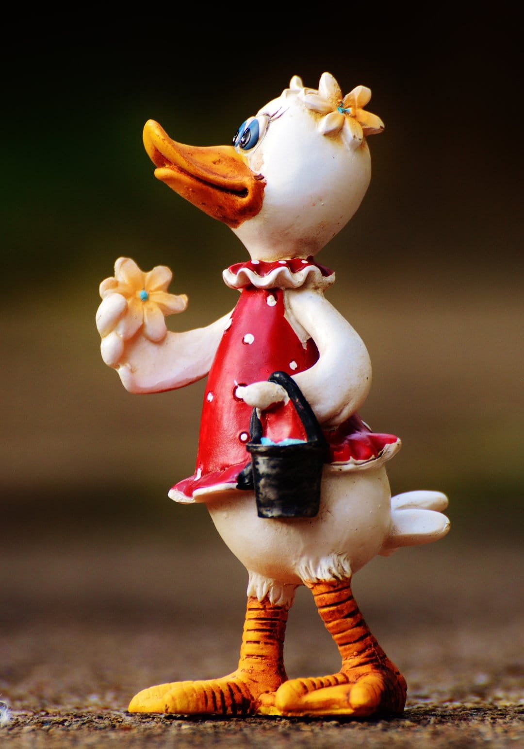 White ceramic duck figurine in a red polka dot dress holding a yellow flower against a blurred background.