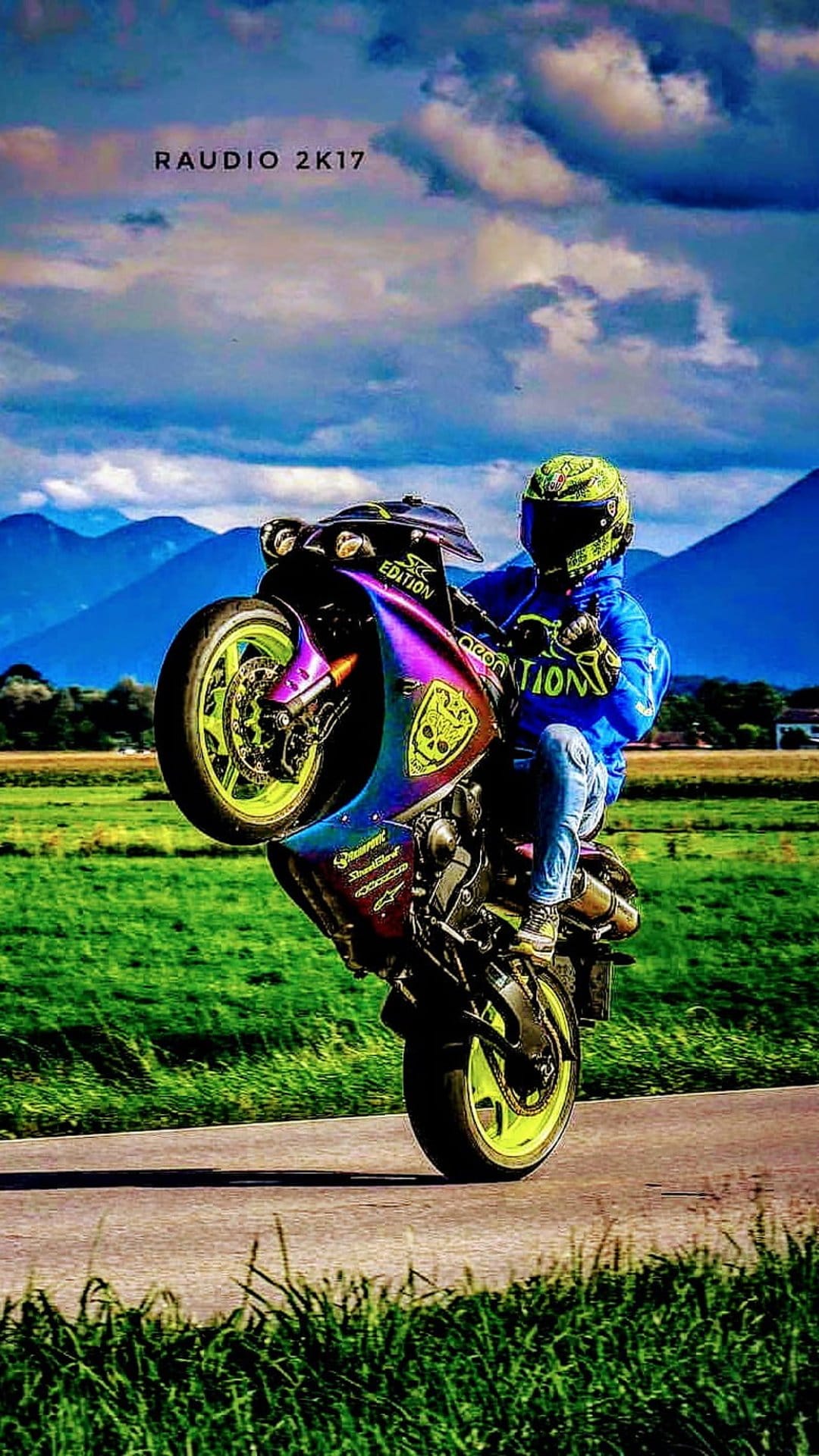 Motorcyclist in a green helmet performs a wheelie on a custom skull bike with rolling hills and mountains behind.