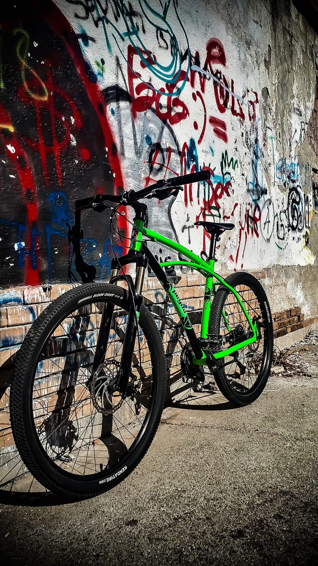 Green mountain bike with detailed spokes parked in front of a colorful, multi-layered graffiti street art wall.