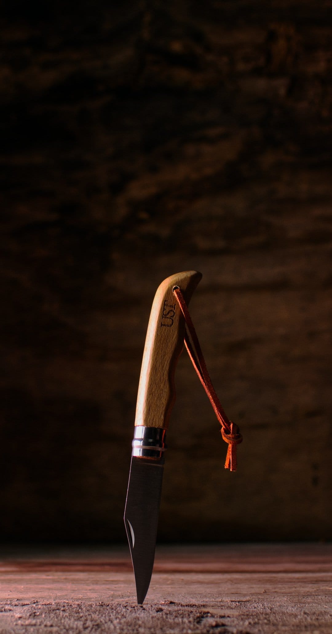 UST folding knife with a wood handle and orange lanyard stands upright on a dark, rustic wooden surface.