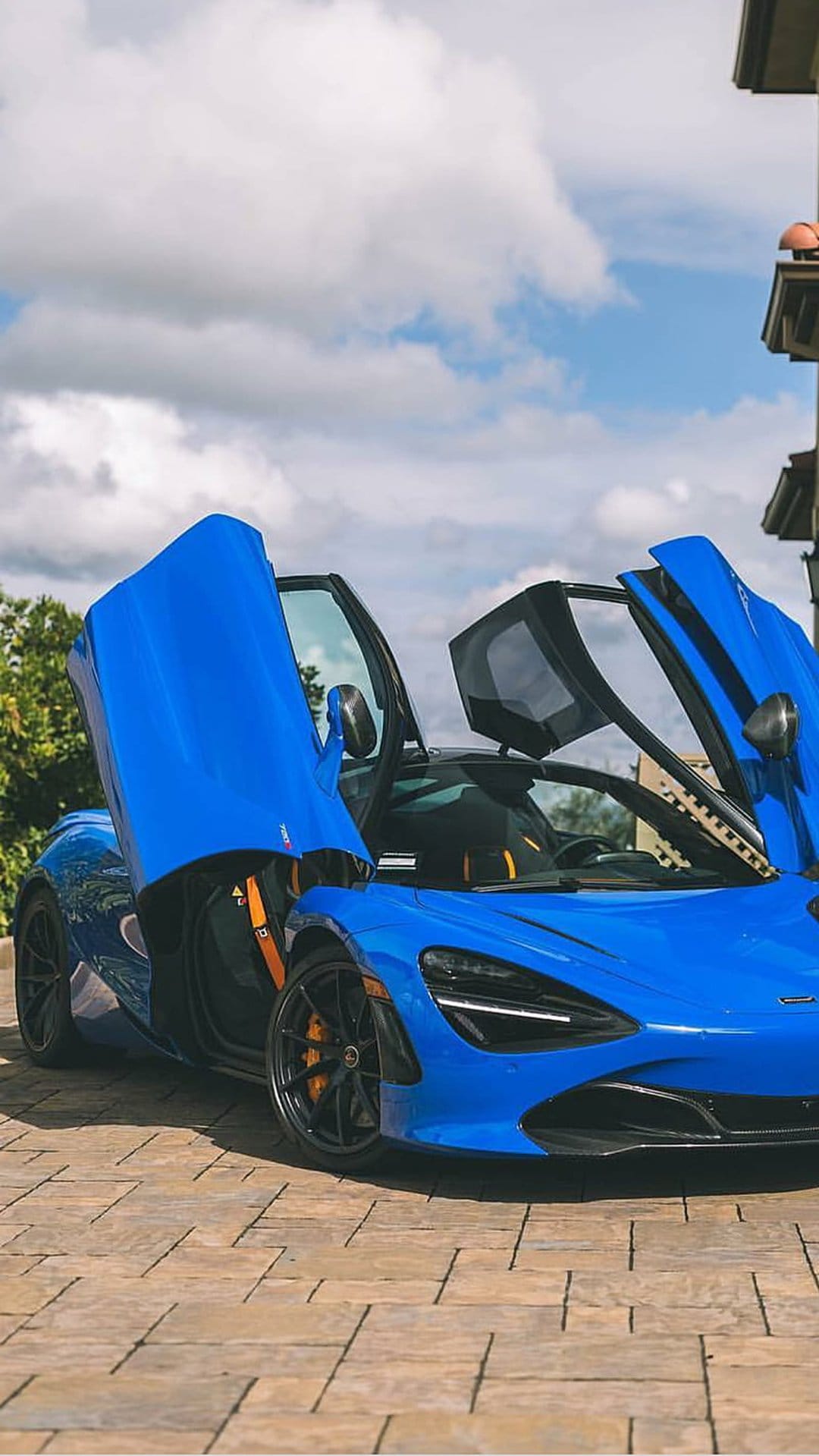 Blue McLaren 720S with open butterfly doors and orange brake calipers parked on a stone driveway outdoors.