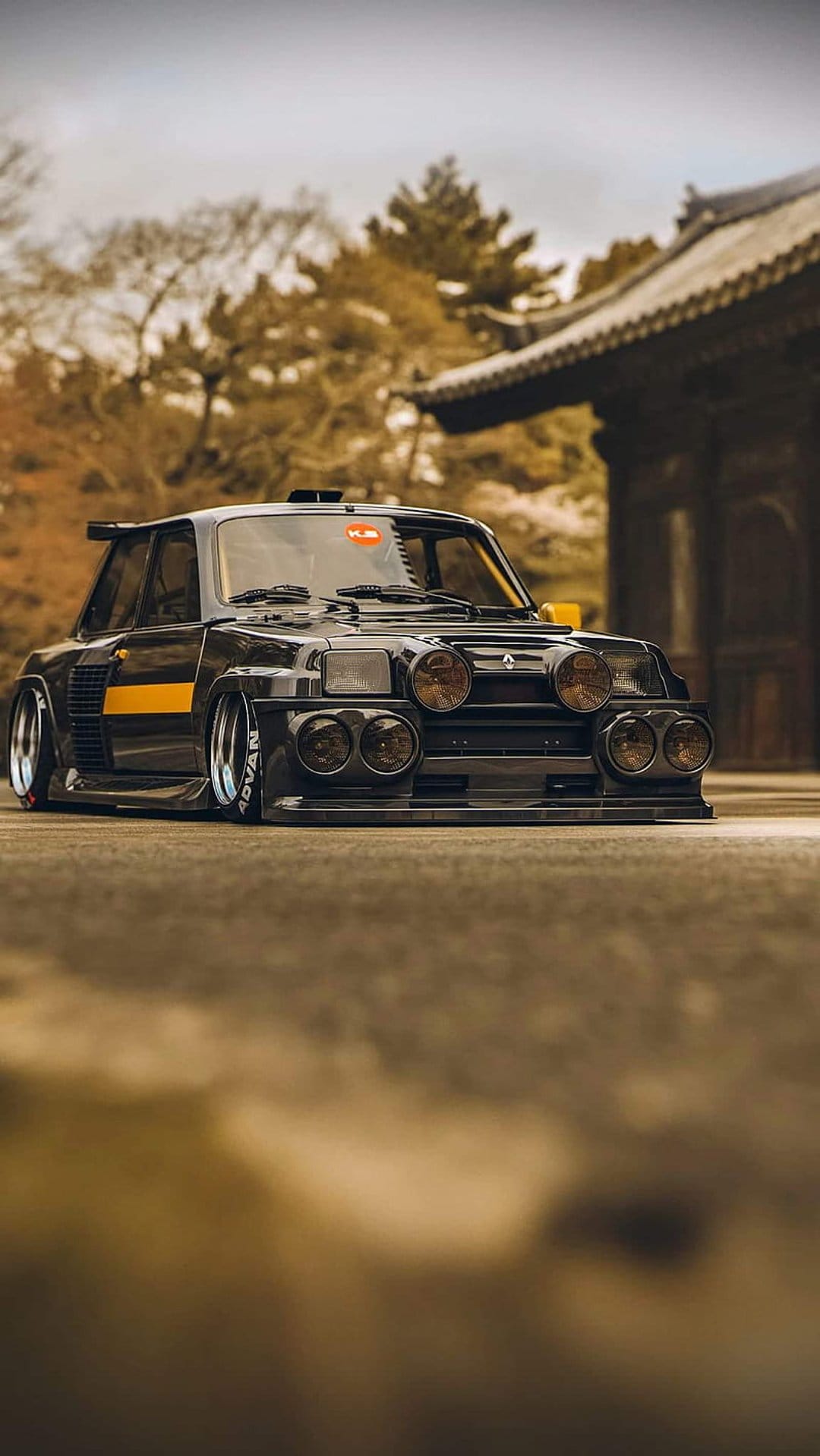 Black Renault 5 Turbo with yellow racing stripes and rally lights parked against Japanese autumn scenery.