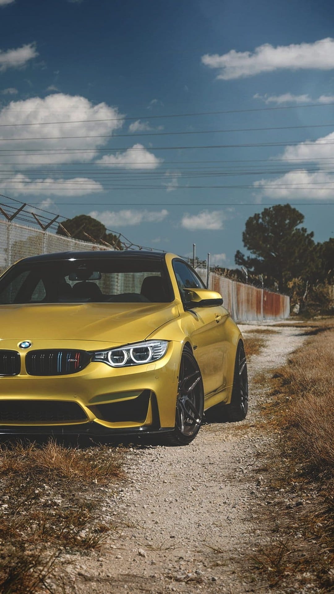 Yellow BMW M4 Coupe with a large kidney grille parked on a gravel road under a cloudy sky near a wire fence.