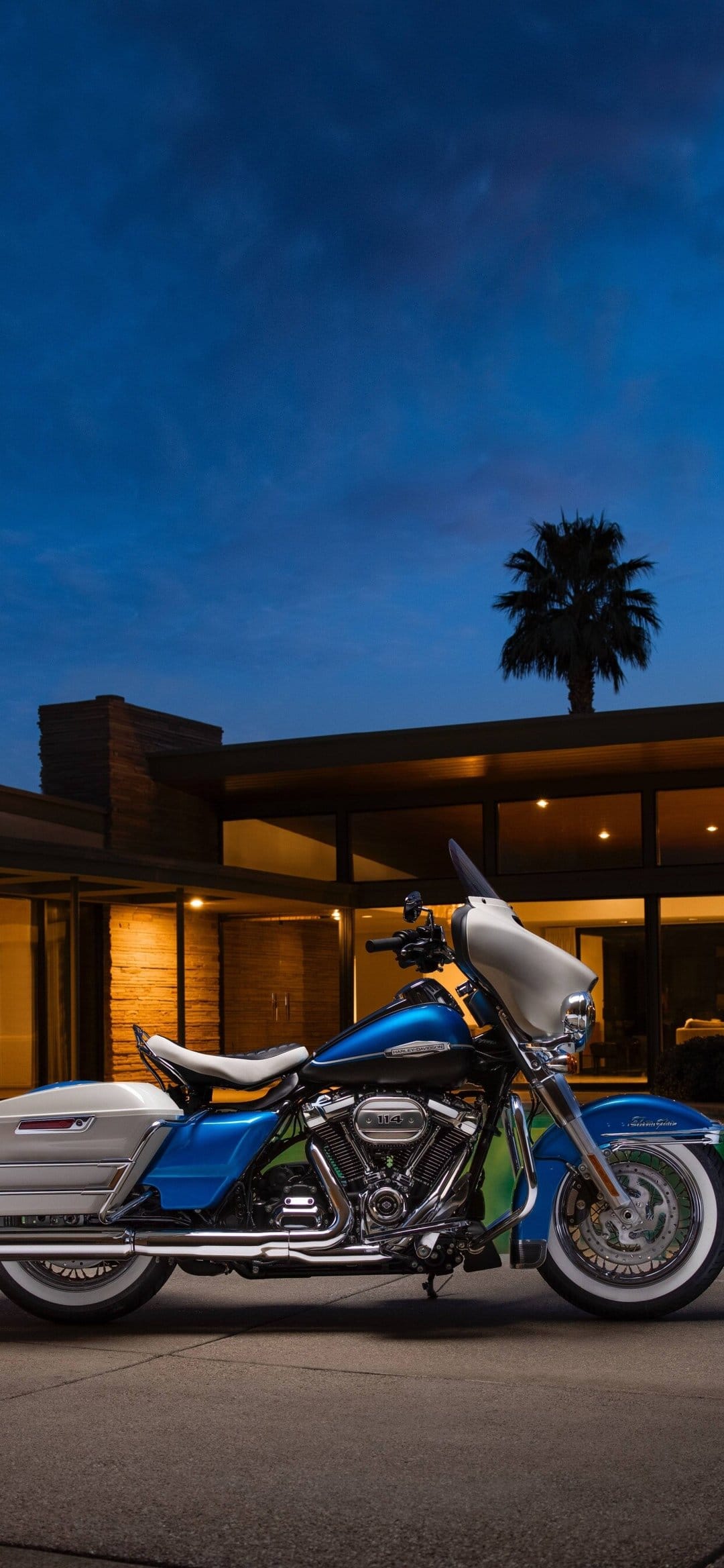 Blue and white Harley-Davidson motorcycle with whitewall tires parked outside a modern house at twilight.