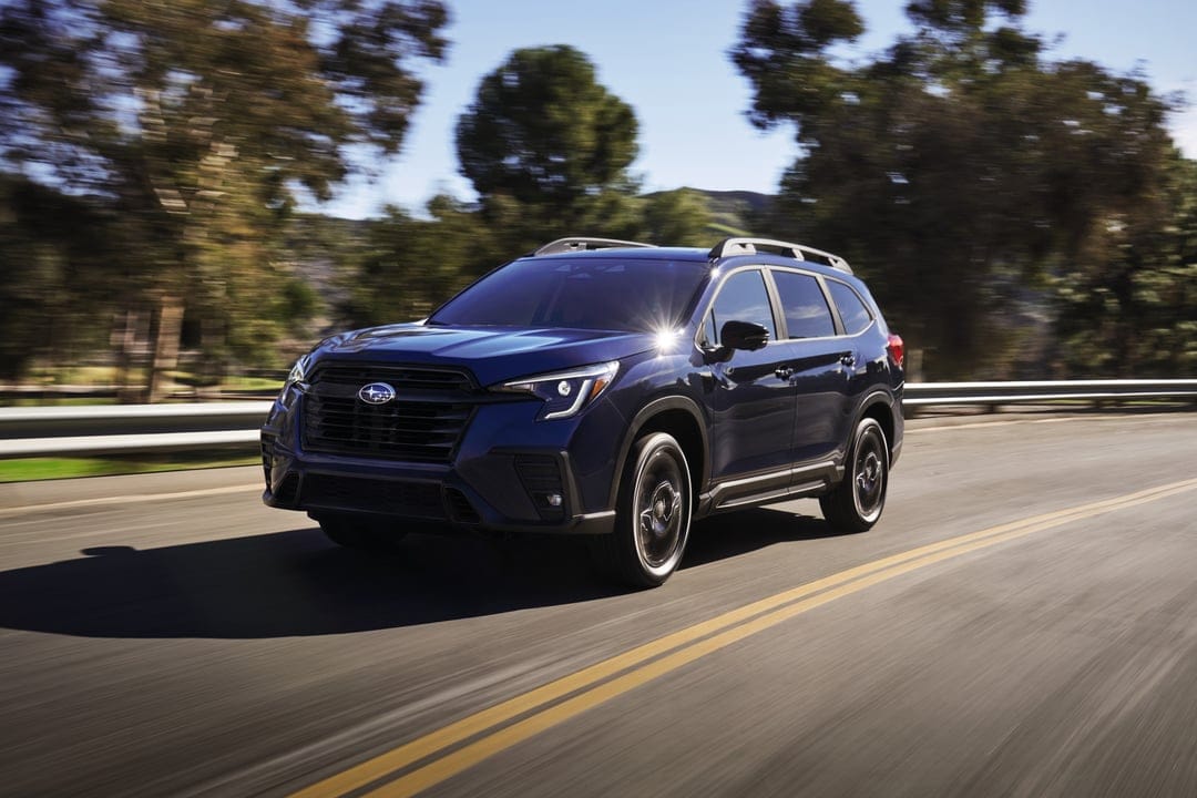 Dark blue Subaru Ascent SUV driving on a sunlit road with motion blur and green forest background.