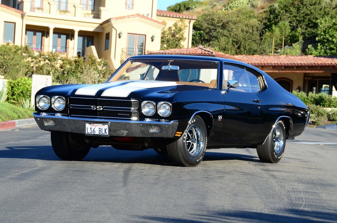 Black 1970 Chevrolet Chevelle SS with white stripes parked on a street near a luxury house and green hills.