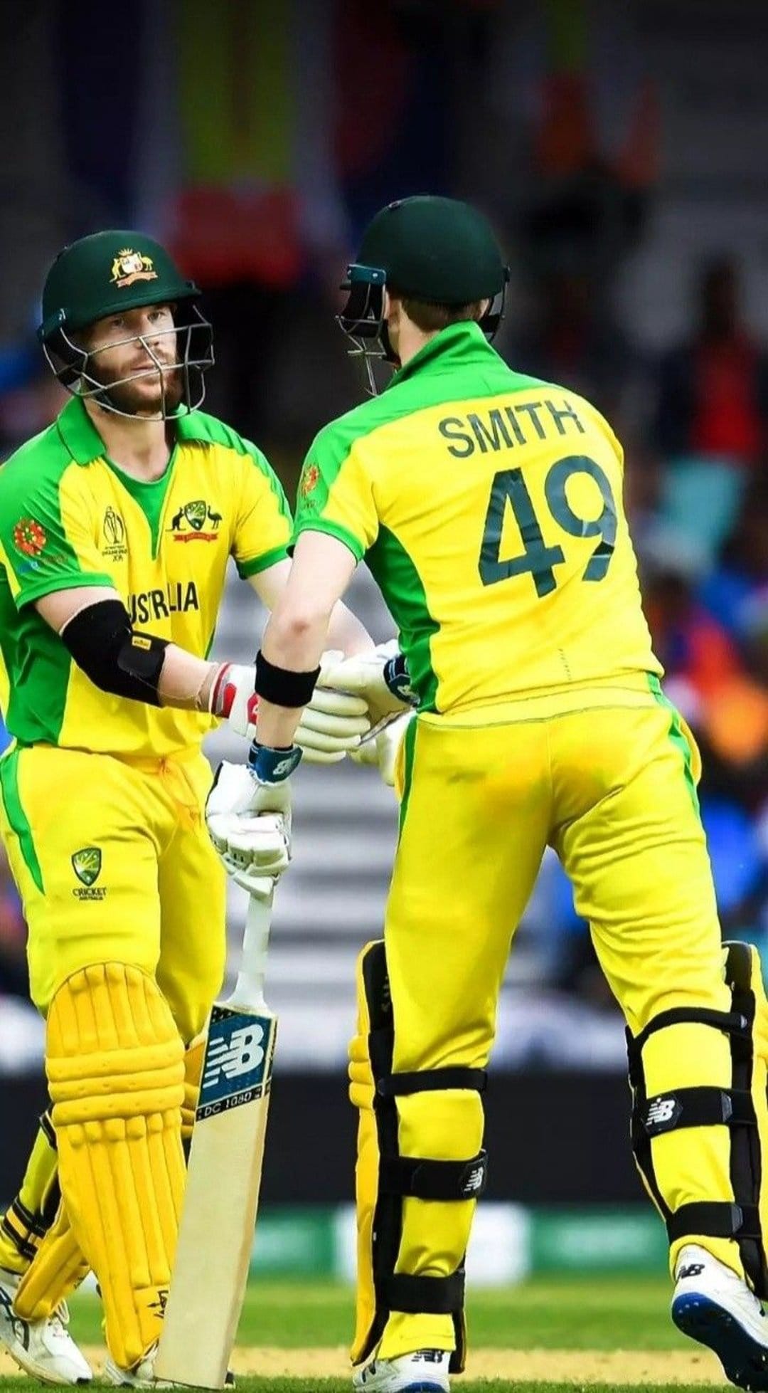 Two cricketers in yellow uniforms shake hands on a stadium pitch while one athlete holds a batting helmet and bat.