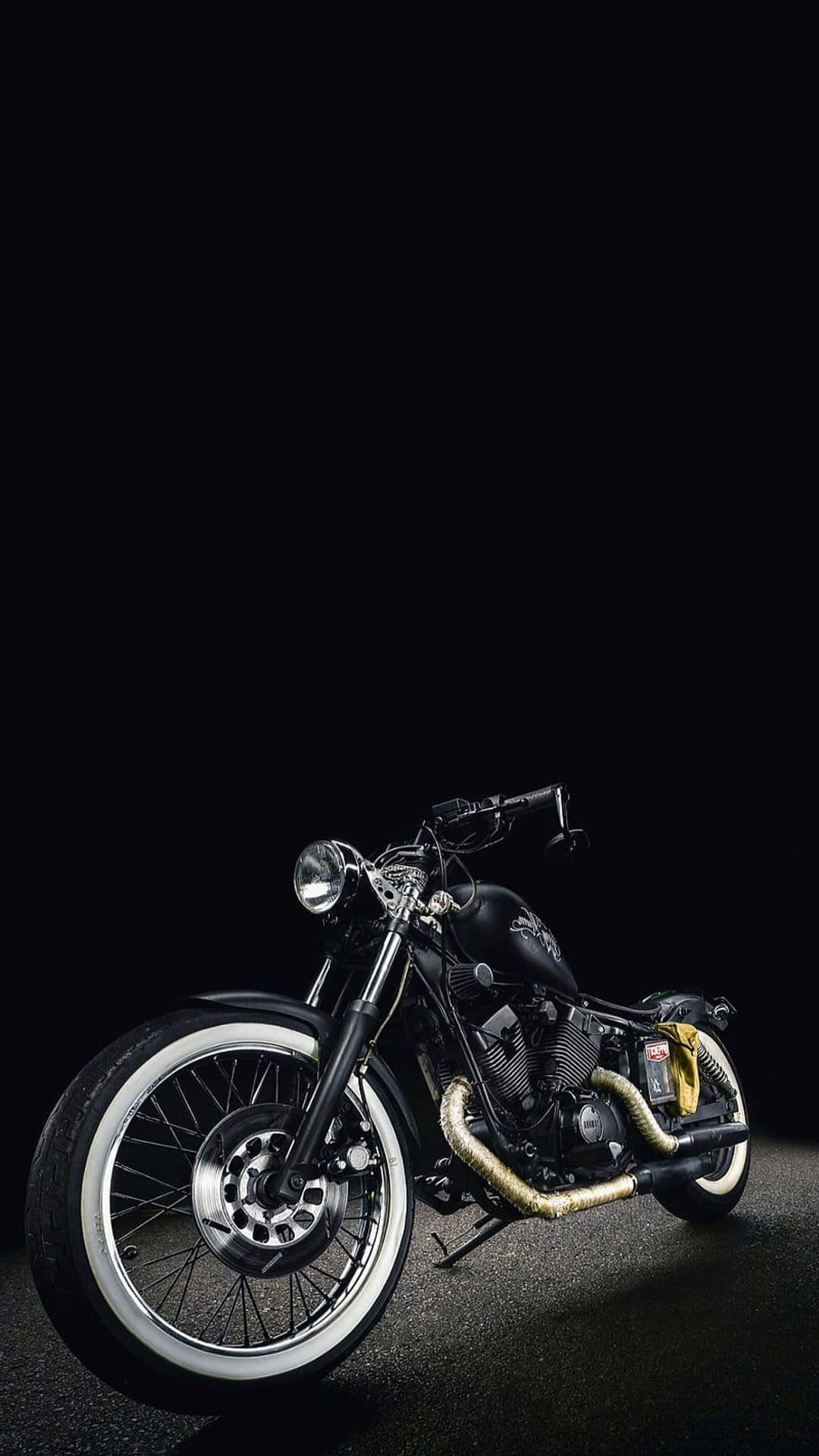 Black cruiser motorcycle featuring gold exhaust pipes and whitewall tires parked in a dark studio setting.