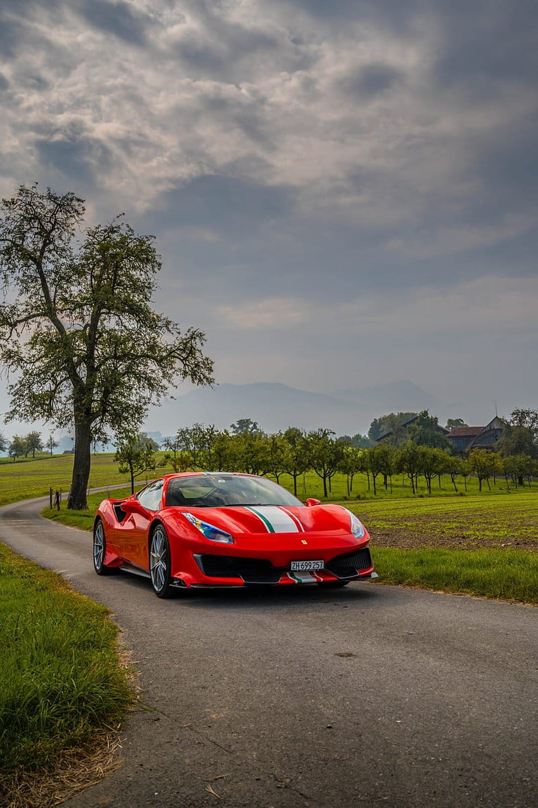 Red Ferrari supercar drives on a winding countryside road with green hills and a cloudy sky in the background.