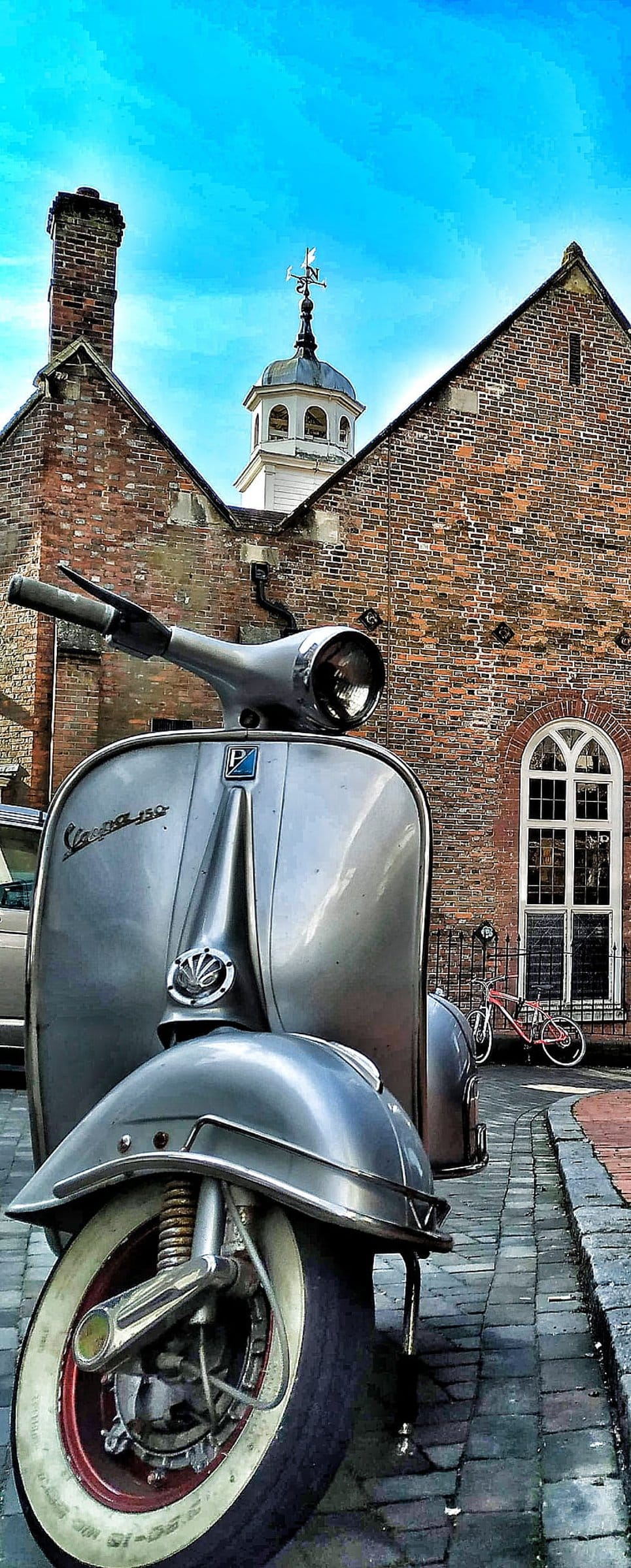 Silver Vespa scooter on cobblestones in front of a red brick tower with an arched window and clear blue sky.