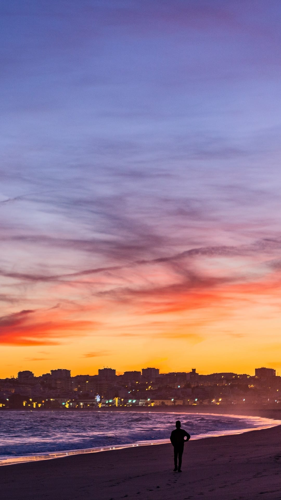 Solitary person walking on a beach at sunset with purple clouds and a distant city skyline in the background.